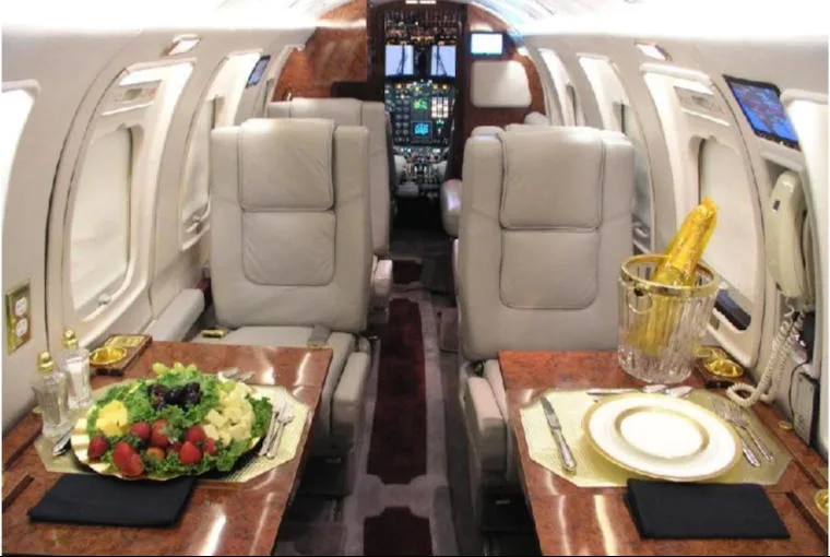 A view of the private jet's cabin interior, featuring light grey seats, wood tables with food and champagne, and a glimpse of the cockpit.