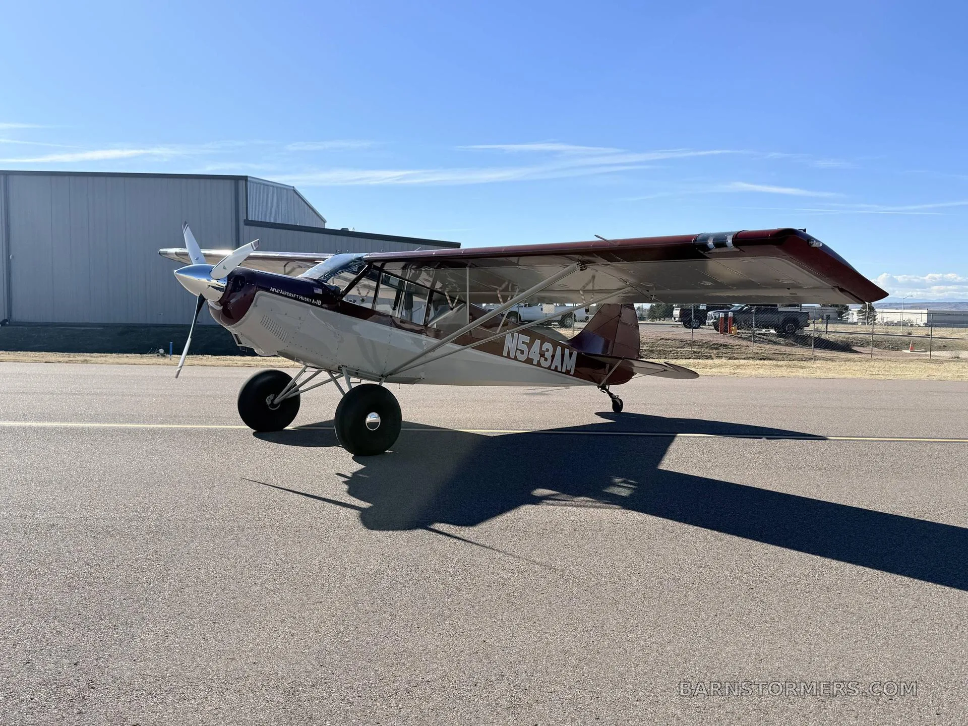 A red and white Aviat Husky A-1B aircraft with large tires is parked on an asphalt runway, viewed from the front-left.