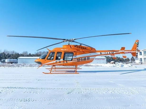 A bright orange helicopter with Chinese text and registration B-728M is parked on a snow-covered field next to buildings.