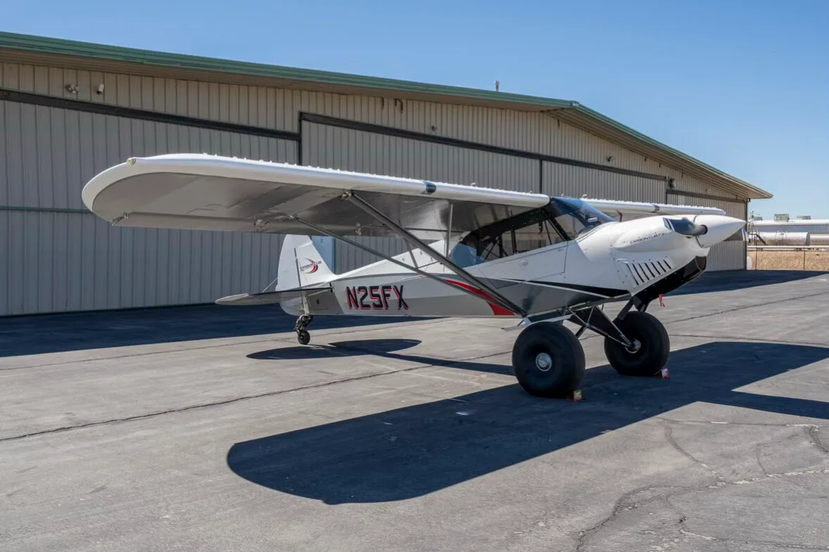 A side view of the white and grey Carbon Cub FX-3 aircraft N25FX parked on an asphalt ramp in front of a hangar.