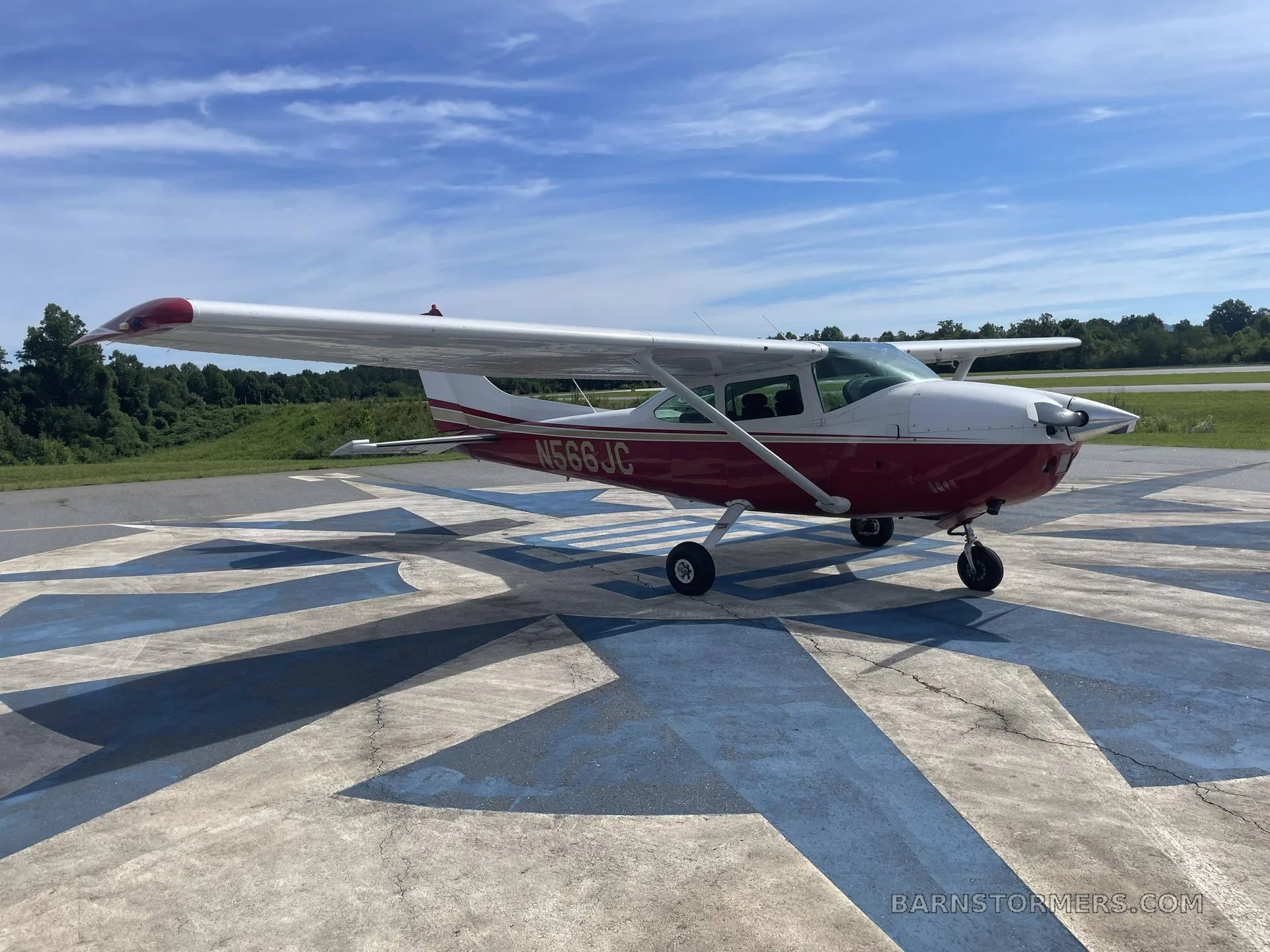 A red and white Cessna N566JC aircraft is parked on a patterned concrete pad at an airfield under a blue sky.