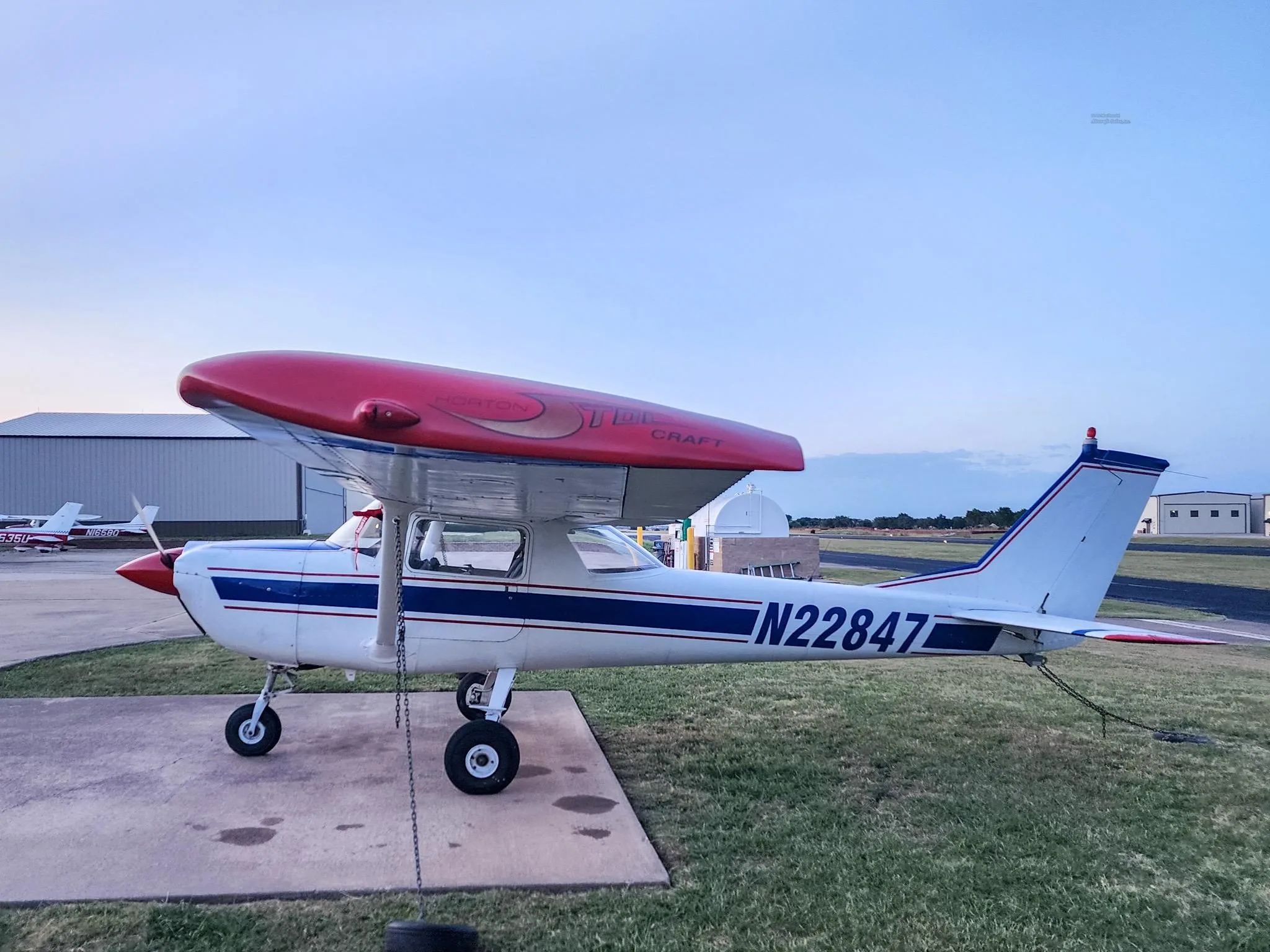 A white Cessna 150 with blue stripes and a red Horton STOL kit on the wing, parked on an airfield at dusk.