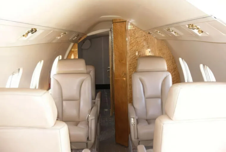 The interior cabin of a private jet, showing rows of beige leather seats and a wooden partition.