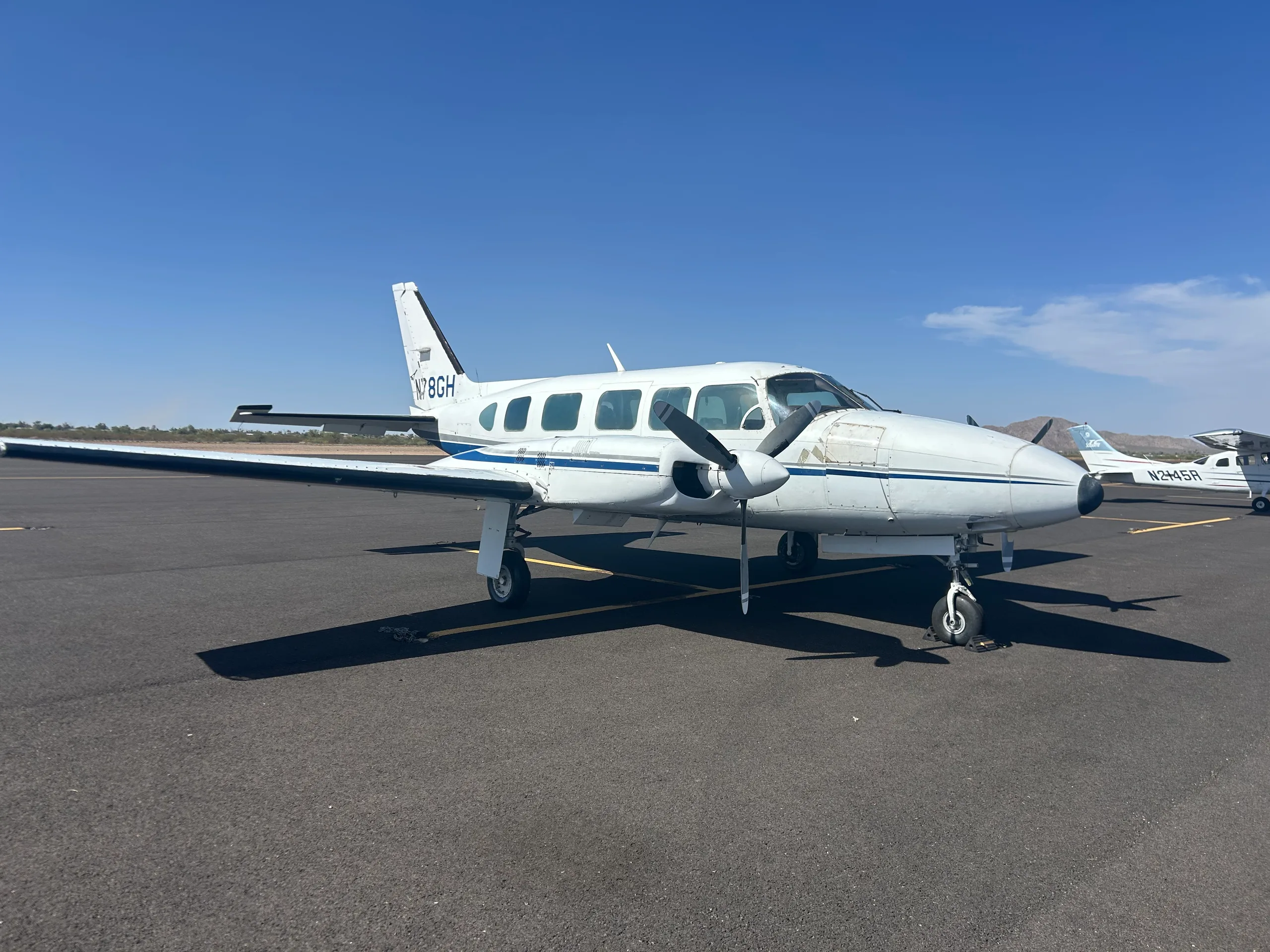 A white twin-engine propeller aircraft with blue stripes, registration N78GH, parked on an asphalt tarmac under a clear blue sky.