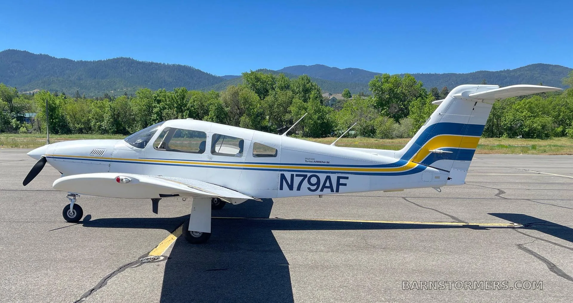 A white Piper Turbo Arrow III aircraft with blue and yellow stripes, registration N79AF, parked on an airfield with mountains in the background.