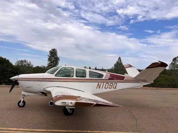 A white Beechcraft Bonanza (N109Q) with brown and maroon stripes is parked on an asphalt surface under a cloudy sky.