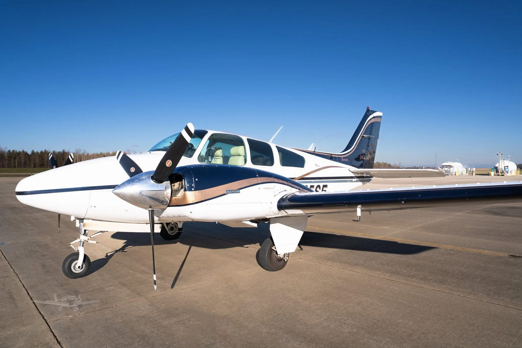 A white and dark blue Beechcraft Baron twin-engine aircraft with tan stripes parked on an airfield under a clear blue sky.
