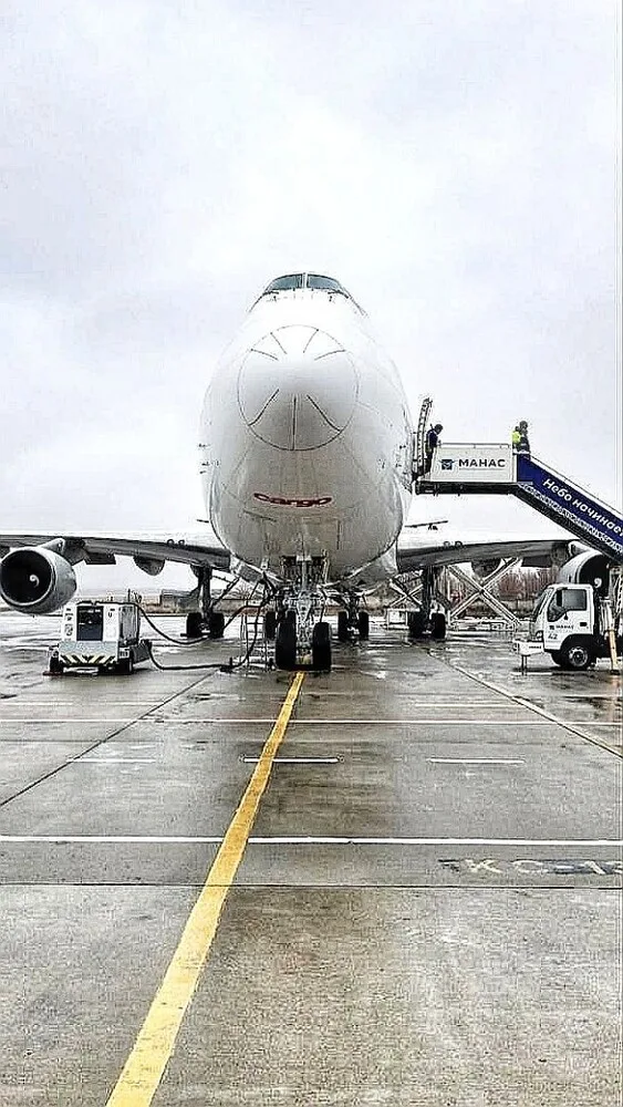A front view of a white Boeing 747 cargo aircraft on a wet tarmac with ground crew and equipment.