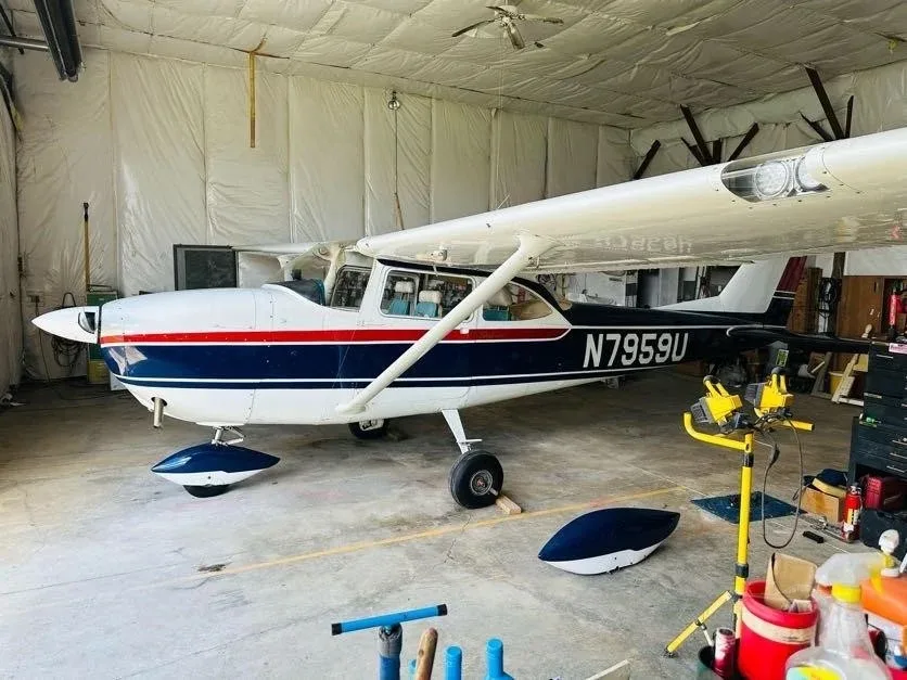 A white and blue Cessna N7959U with a red stripe is parked in a hangar, with a wheel fairing removed.