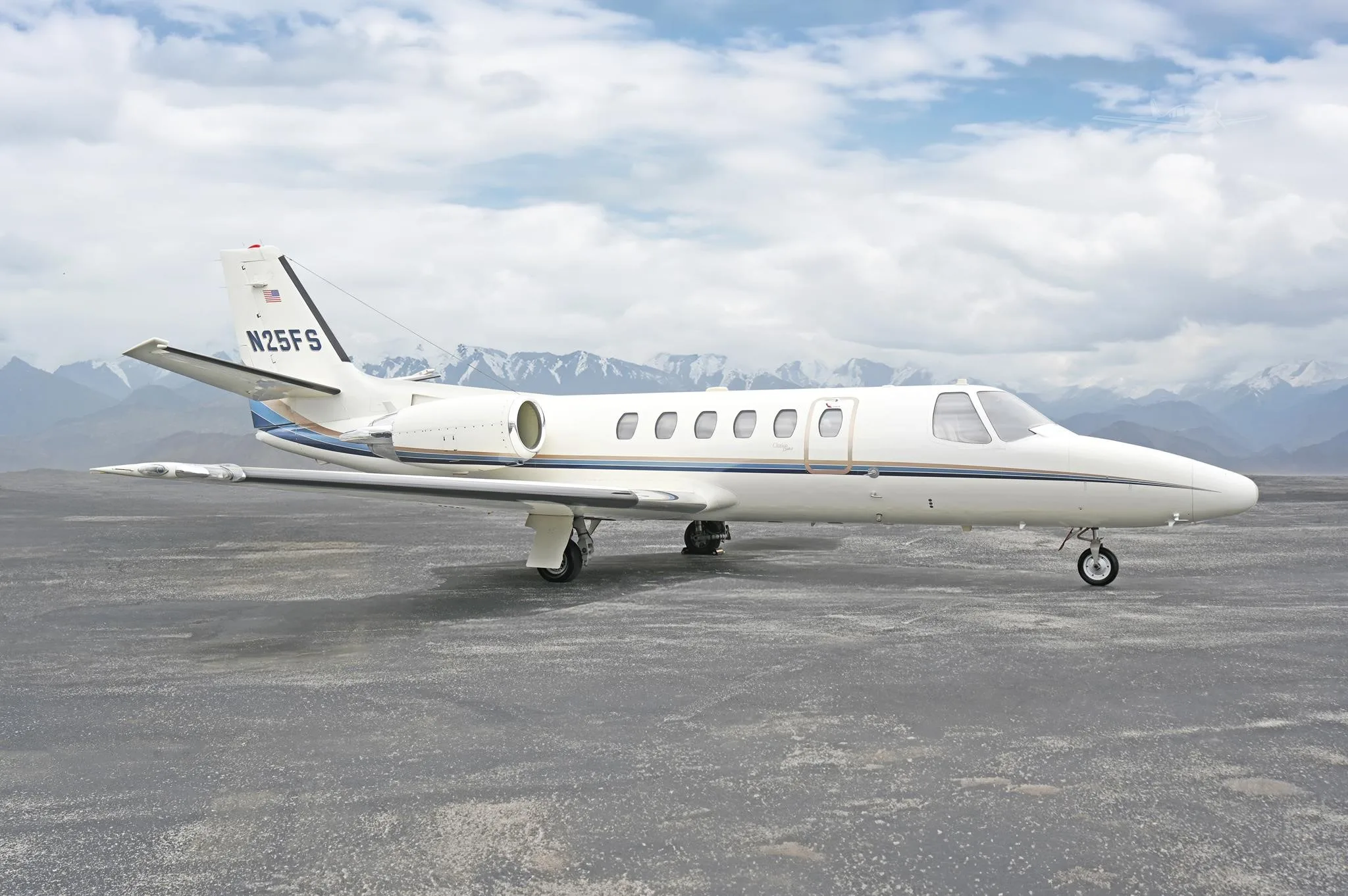 A white Cessna Citation business jet with blue and gold stripes is parked on a tarmac with mountains in the background.