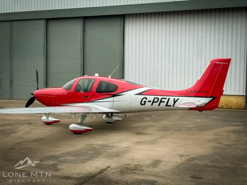 A red and white Cirrus SR22 aircraft (G-PFLY) parked in profile on a tarmac in front of a hangar.