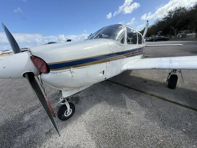 A front-side view of the white Piper Cherokee aircraft, showing its propeller, landing gear, and blue and red stripes.