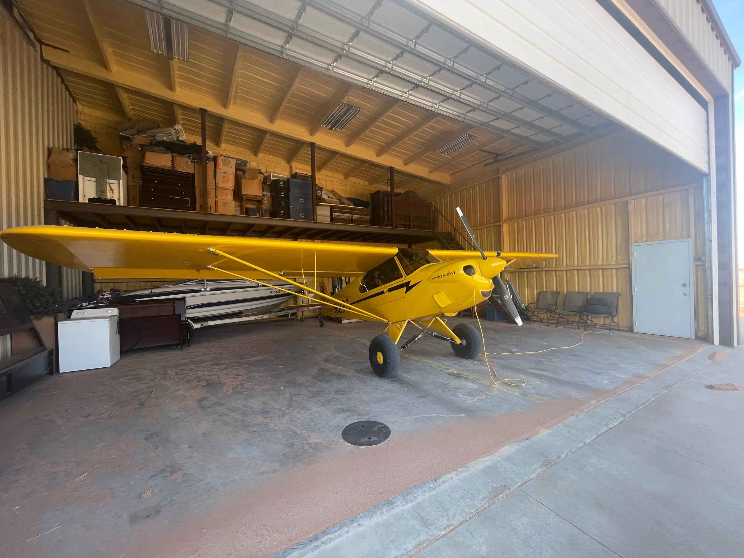 A yellow Sport Cub S2 aircraft with black stripes is parked inside a hangar, with a boat and other storage items in the background.