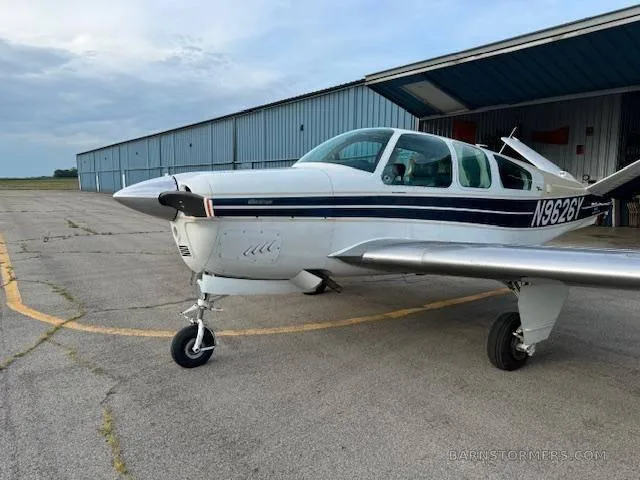 A front-side view of a white and blue Beechcraft Bonanza with registration N9626Y, parked on an airfield next to a blue hangar.