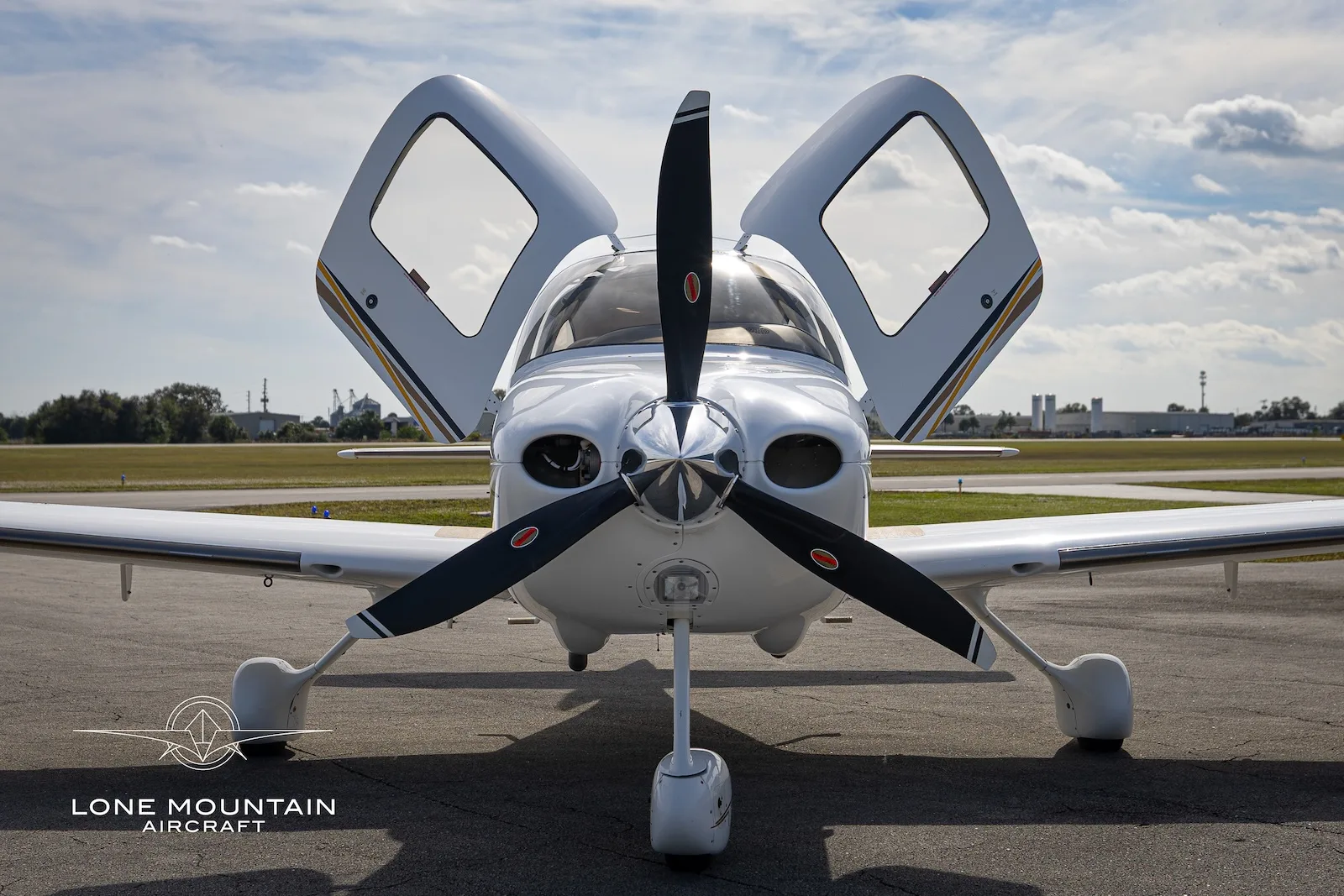 A head-on view of a white Cirrus SR22 aircraft with gold and black accents, registration N323CD, parked on an airfield with its doors open.