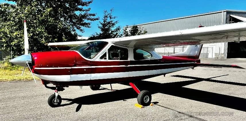 A red and white single-engine high-wing aircraft parked on an asphalt ramp in front of a hangar.