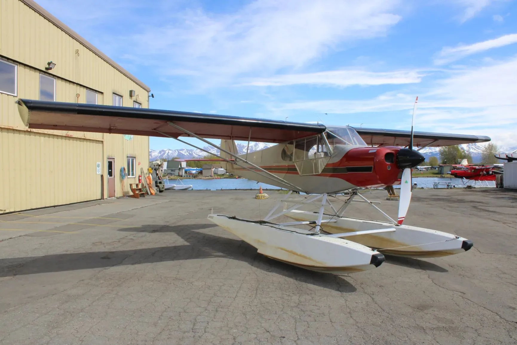 A red and grey floatplane parked on an asphalt apron next to a yellow hangar, with mountains and water in the background.