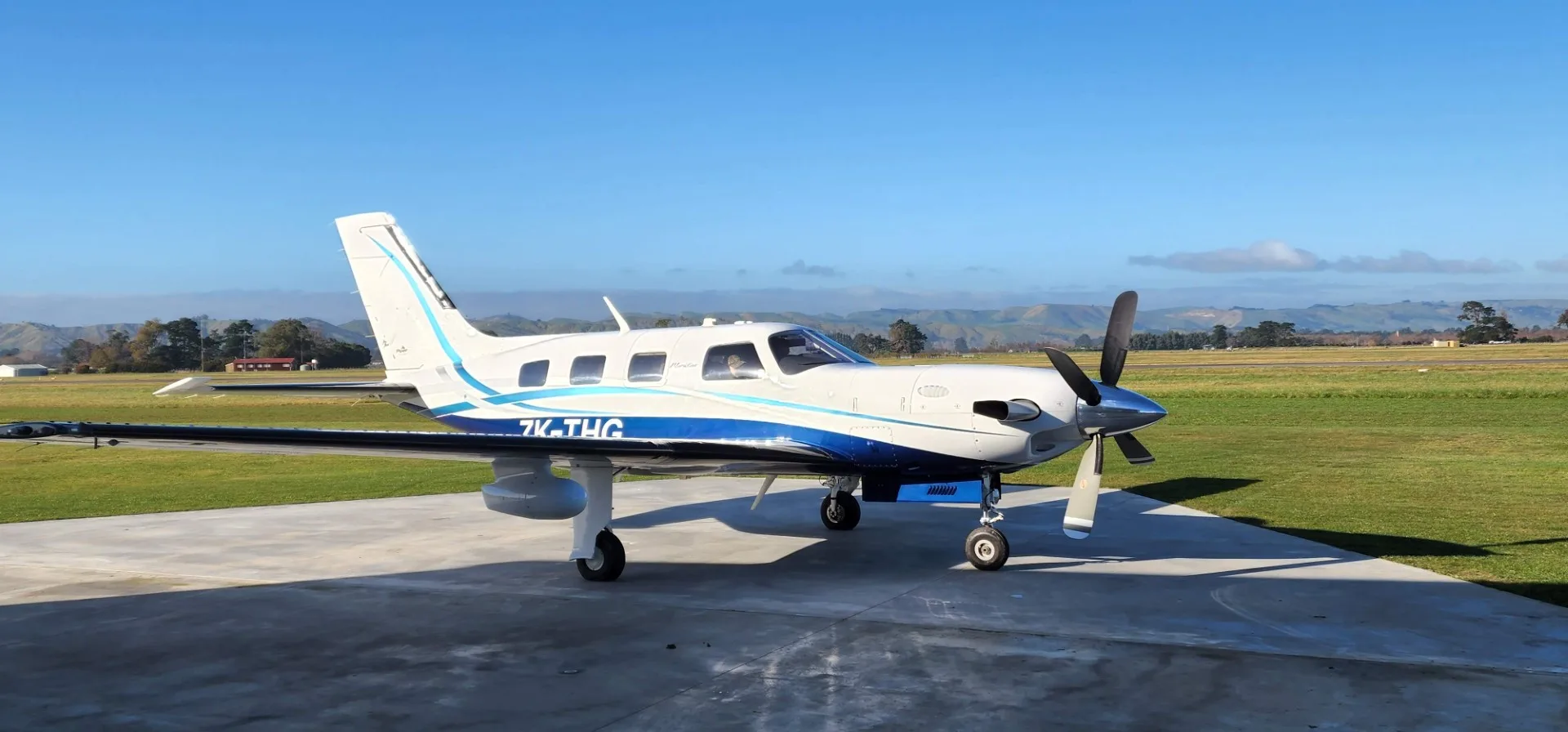 A white and blue Piper M600 aircraft, registration ZK-THG, parked on a concrete pad at an airfield under a clear blue sky.