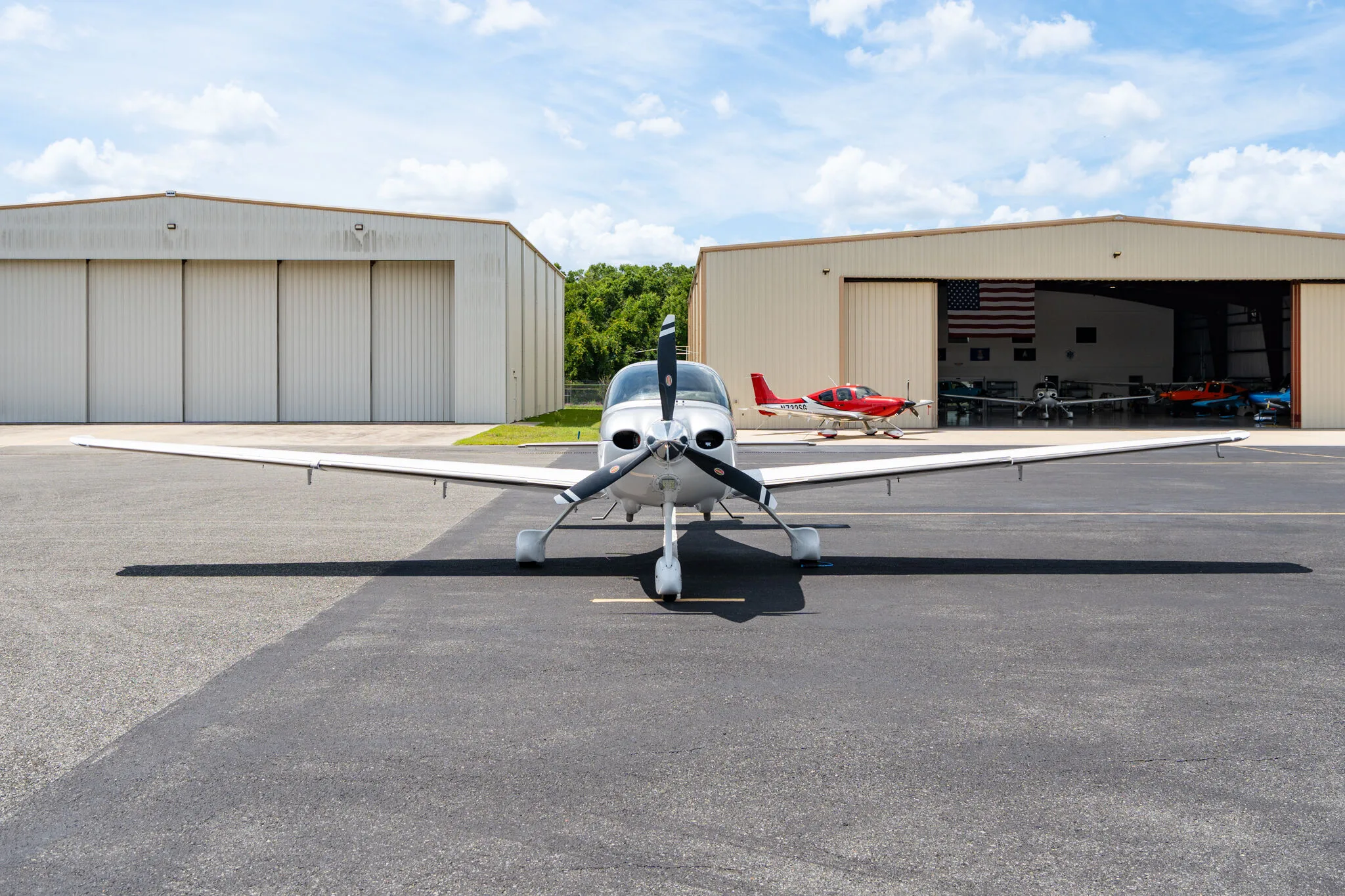 Front view of a silver Cirrus SR22 aircraft parked on an airfield with hangars in the background.