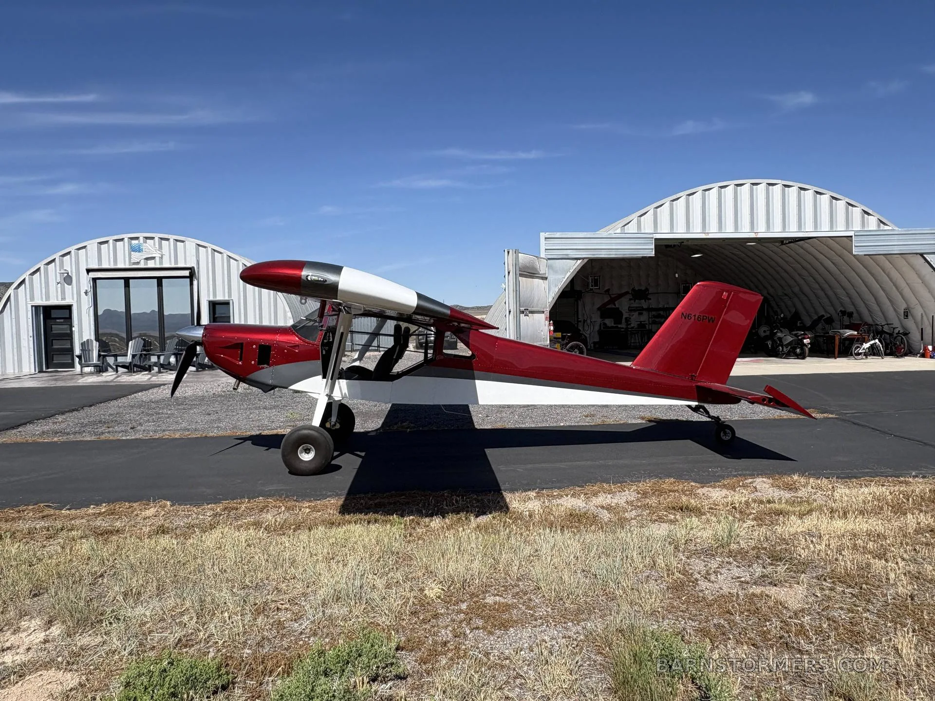 A red and white single-engine aircraft with tail number N616PW parked on an asphalt strip in front of two hangars.