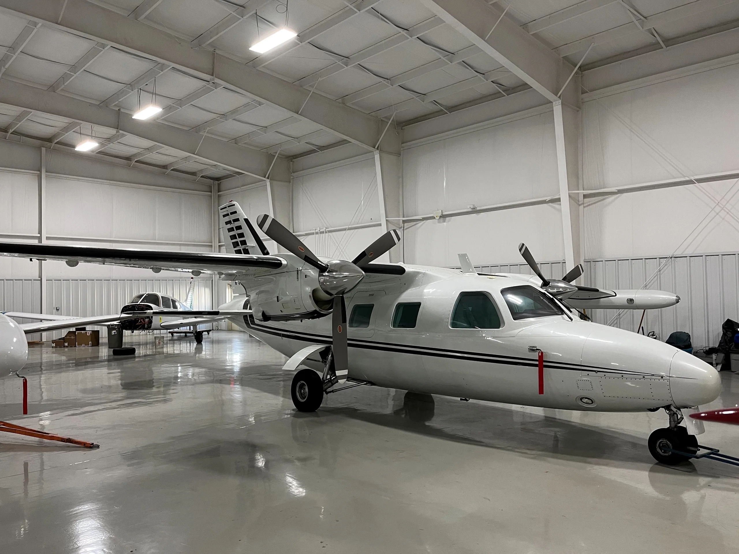 A white twin-engine turboprop aircraft with black stripes is parked in a brightly lit hangar, with another smaller aircraft visible in the background.