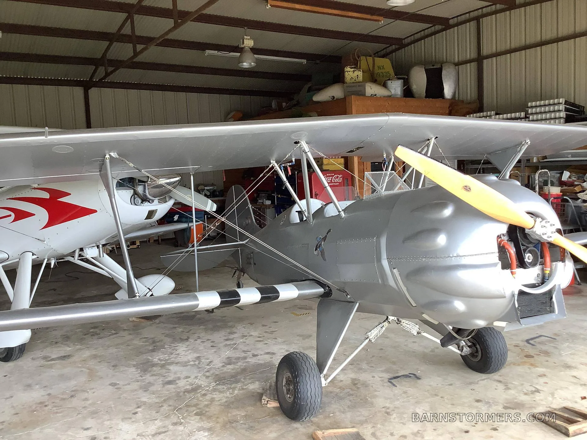 A silver biplane with a wooden propeller is parked in a hangar, with another white aircraft partially visible behind it.