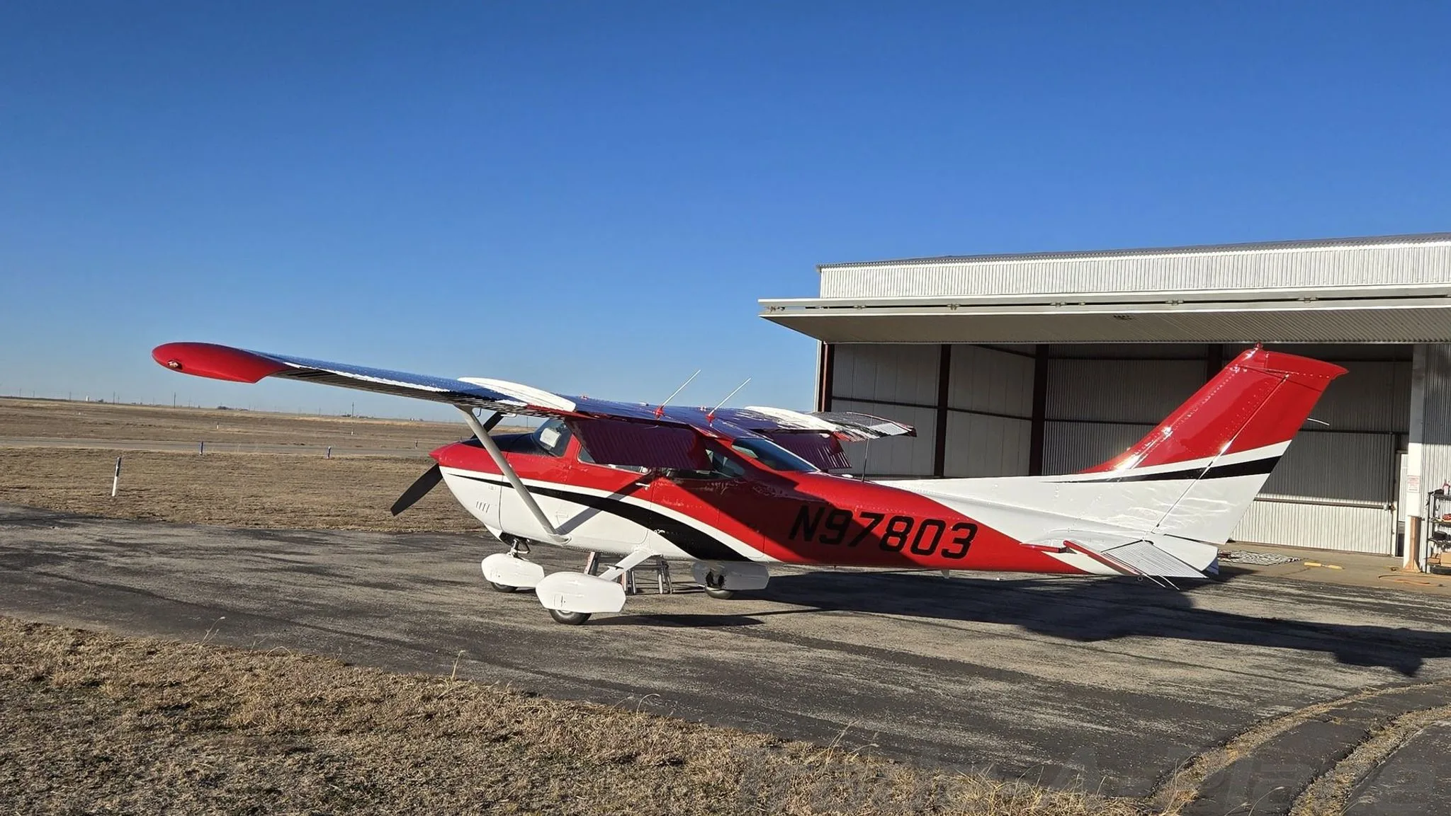 A red, white, and black Cessna aircraft (N97803) is parked on an airfield under a clear blue sky.