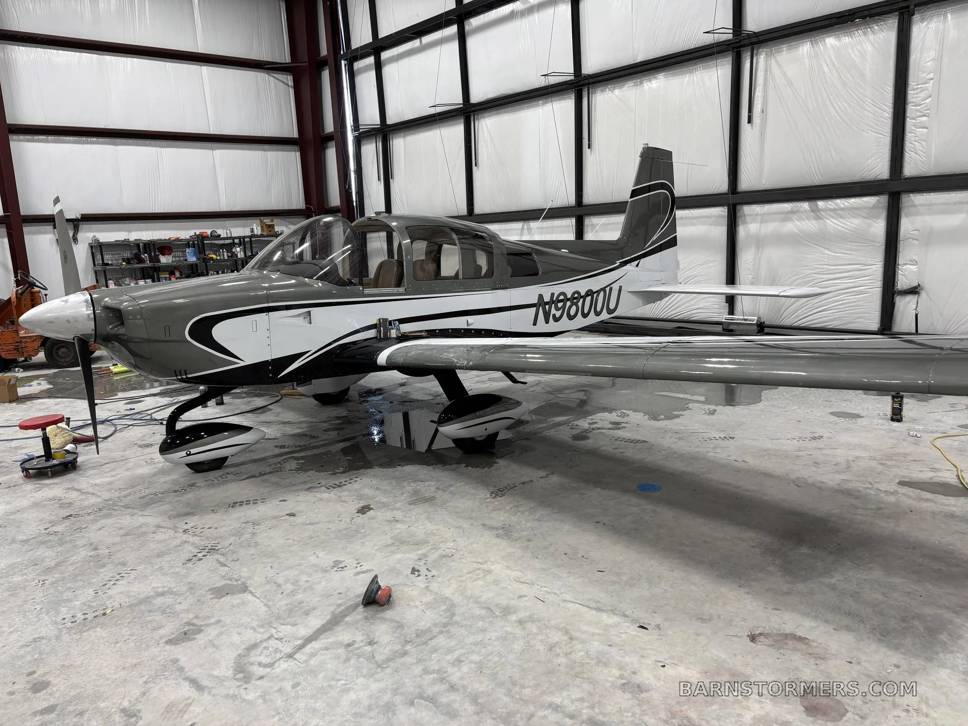 A gray and white Grumman American AA-5B Tiger (N9800U) parked inside a hangar with tools and equipment nearby.