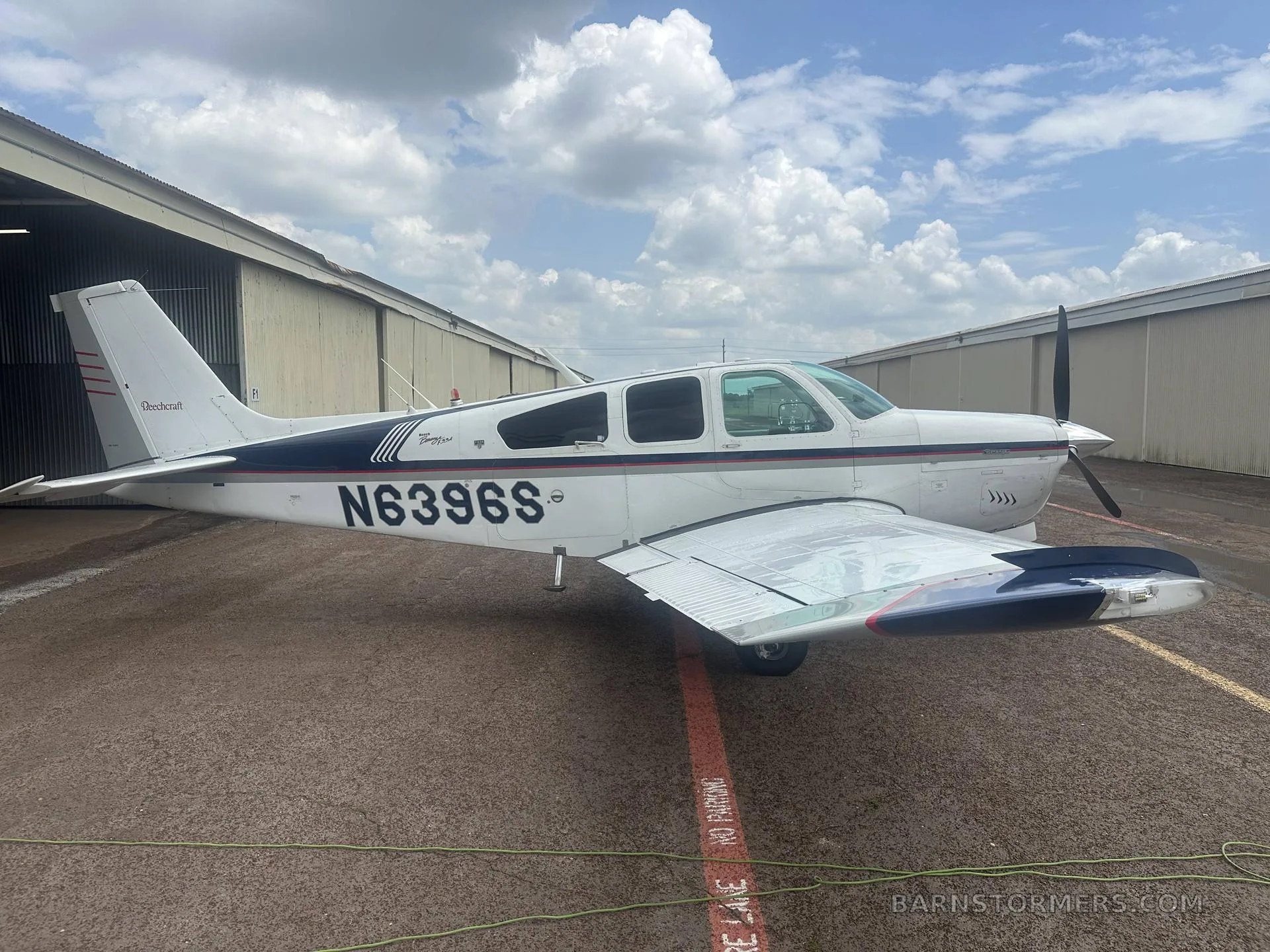 A white and blue Beechcraft Debonair N6396S parked on an asphalt apron next to a hangar under a cloudy sky.