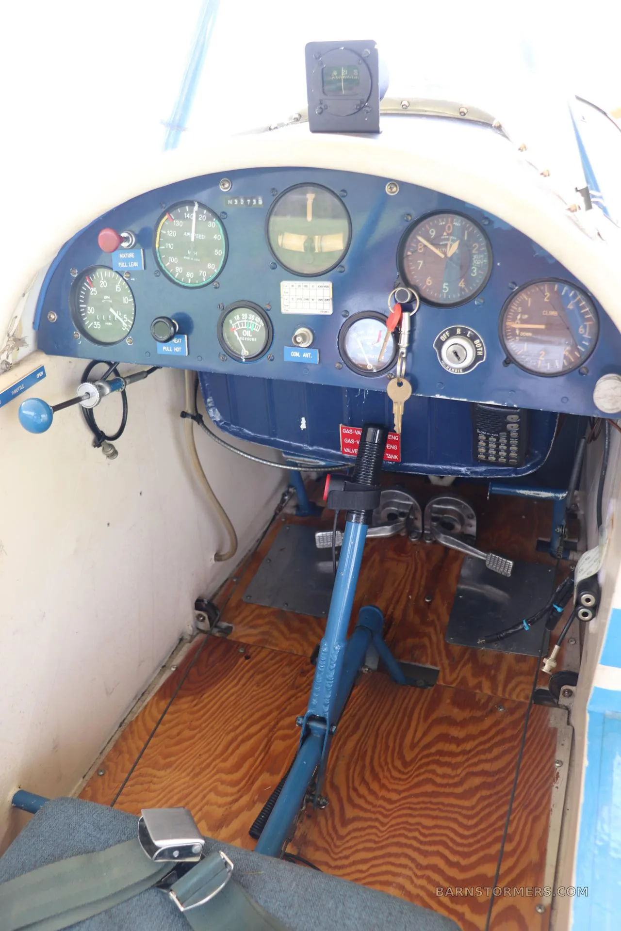 The cockpit of a light aircraft with a blue instrument panel, wooden floorboards, and a single seat.