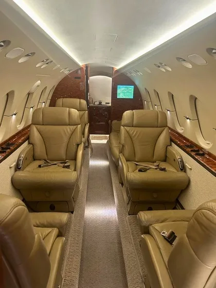 The interior cabin of a business jet, featuring tan leather seats arranged in a club configuration with a central aisle and wood paneling.