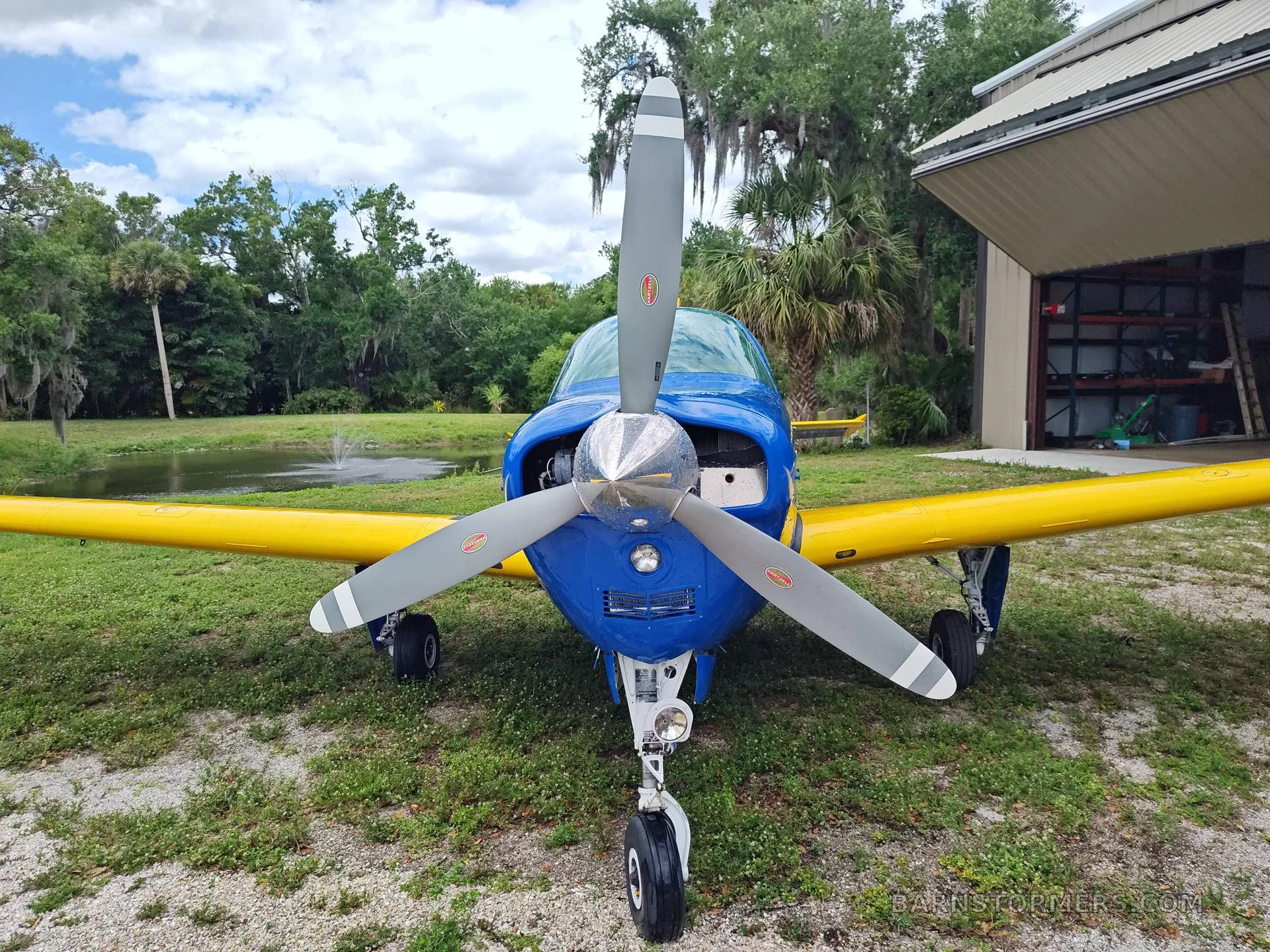 A blue and yellow single-engine aircraft is viewed from the front, showing its propeller and nose gear, parked in front of a hangar and pond.