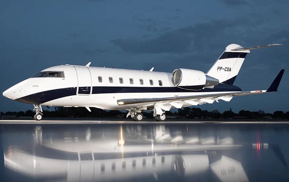 A white and dark blue Bombardier Challenger business jet (PP-COA) parked on a wet tarmac at dusk.
