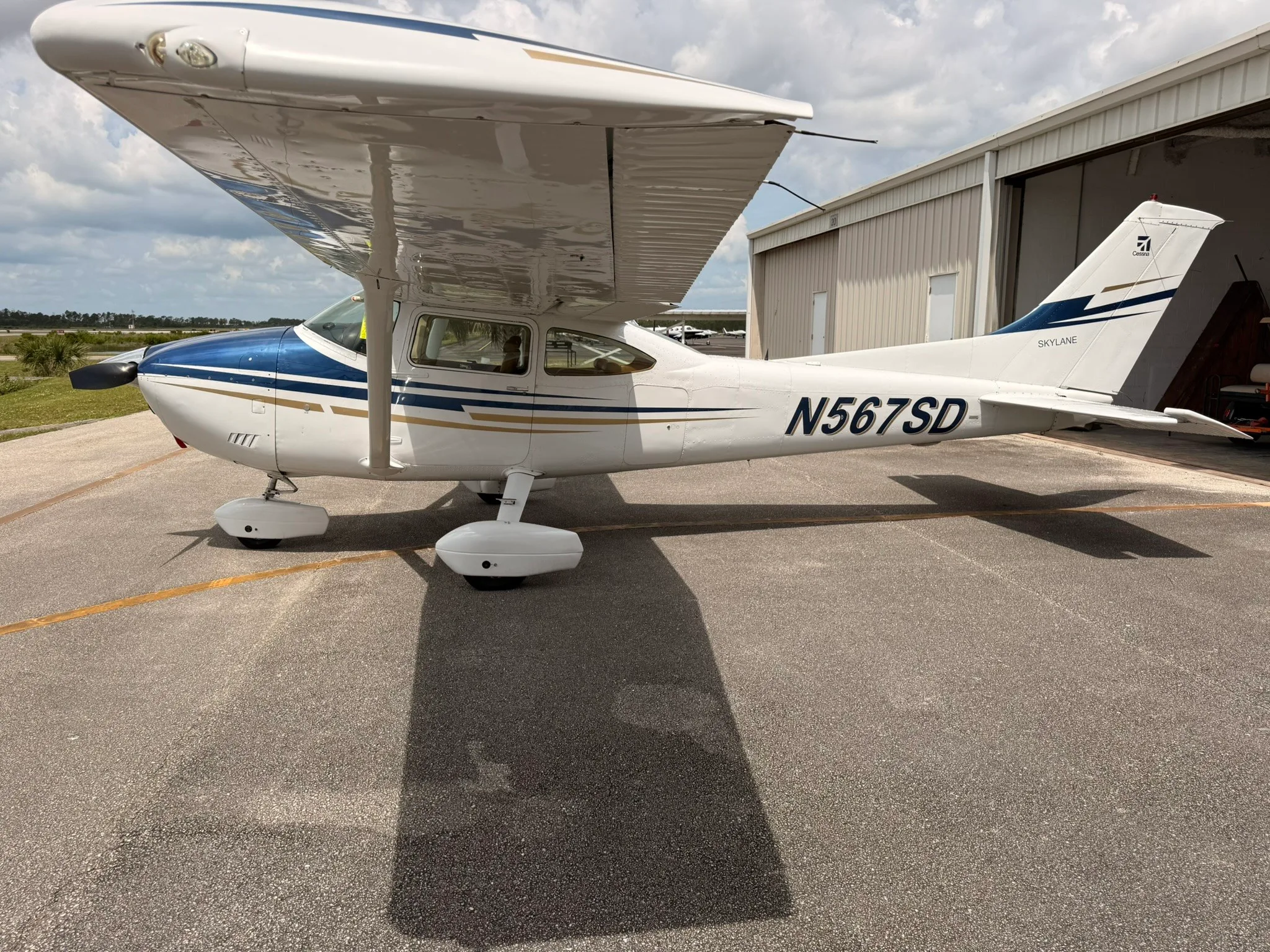 A white Cessna Skylane N567SD with blue and gold stripes parked on an asphalt ramp, viewed from the front-right.