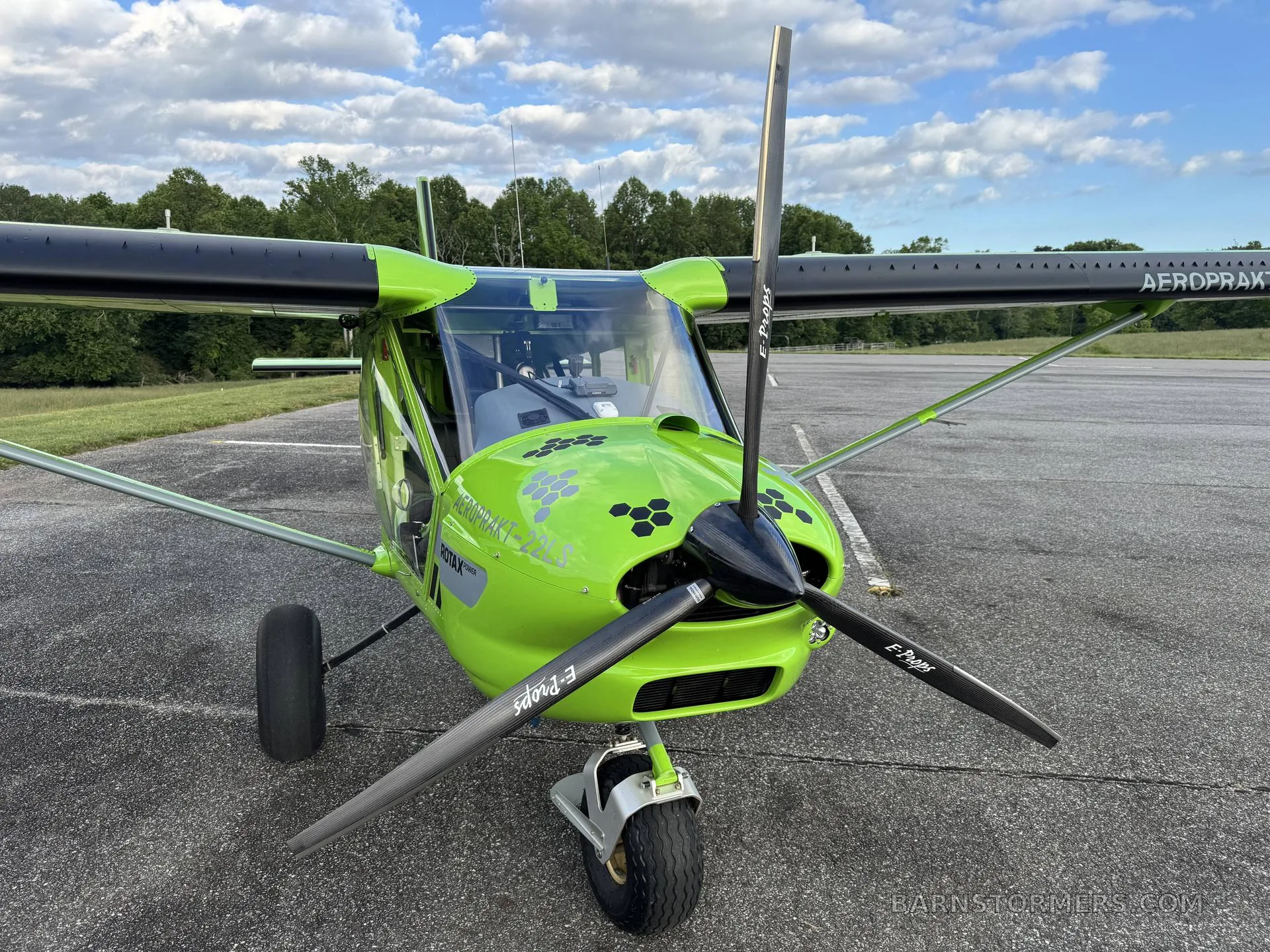 A front view of a bright green Aeroprakt-22LS light sport aircraft with a three-blade propeller, parked on an asphalt runway.