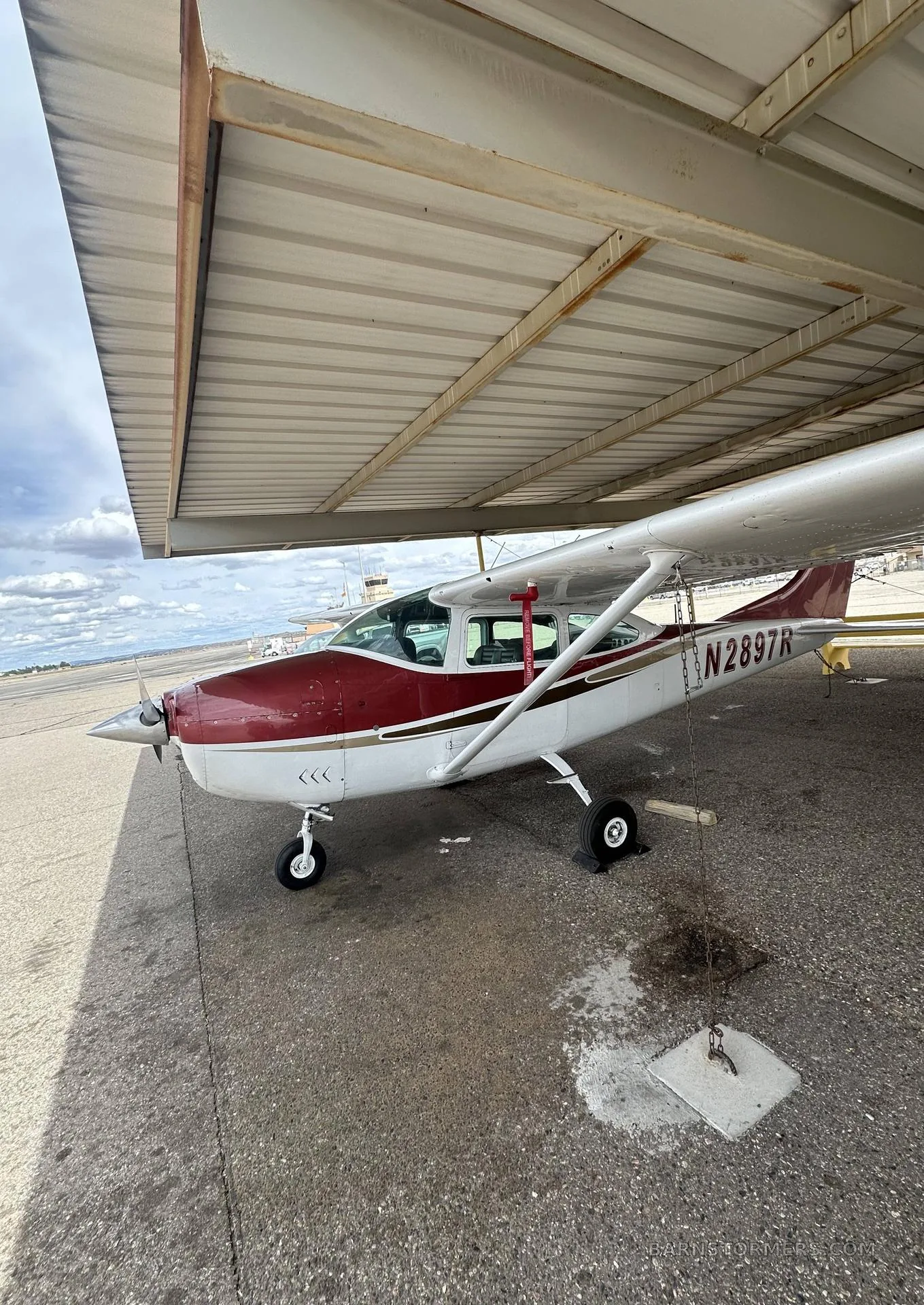 A red and white single-engine propeller aircraft (Cessna N2897R) is parked under a metal shelter on an asphalt ramp.