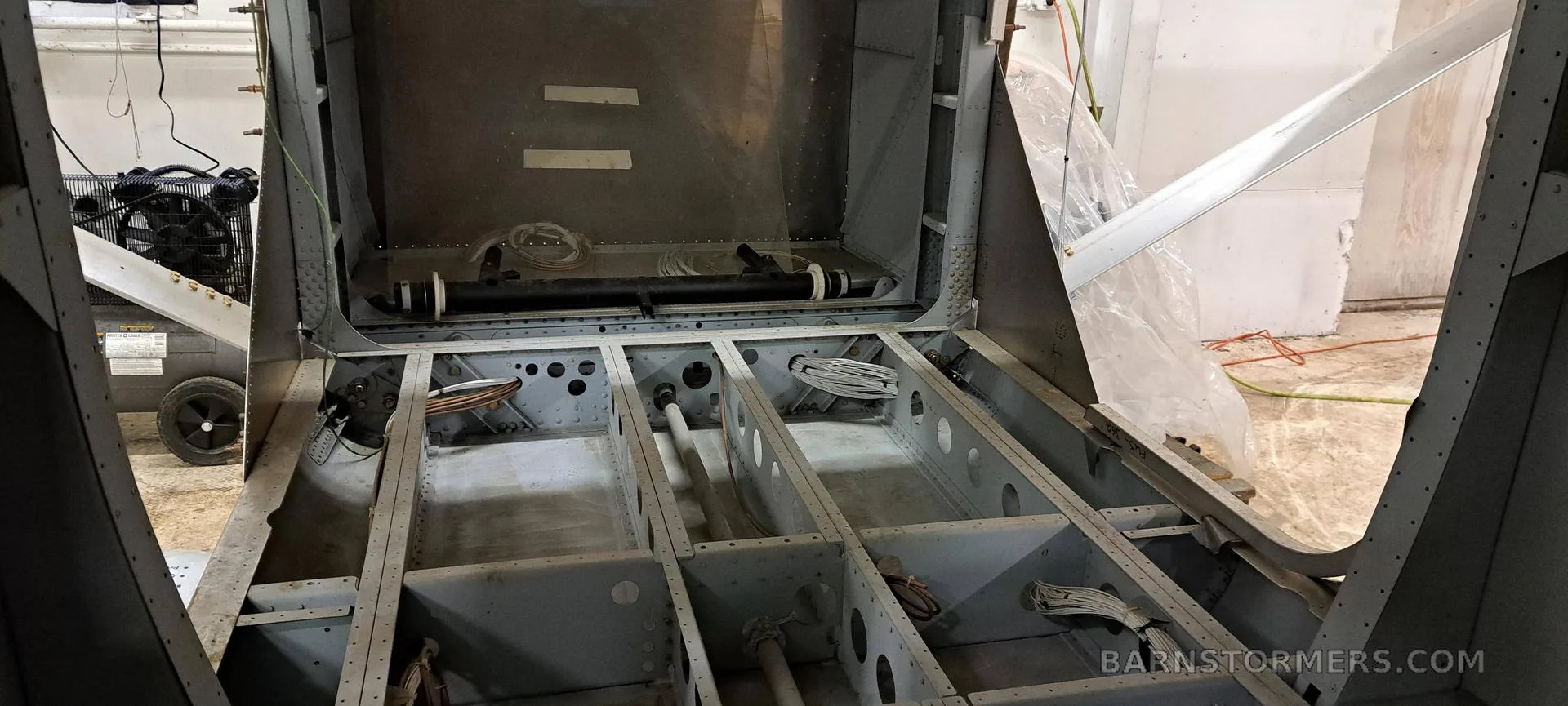An overhead view of an aircraft fuselage frame with internal wiring and structural components visible in a workshop.
