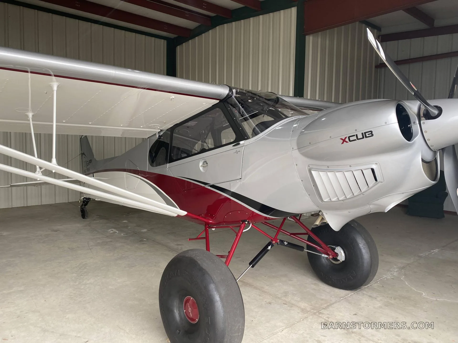 A silver and red X-Cub aircraft with large tundra tires, parked inside a hangar, viewed from the front-right.