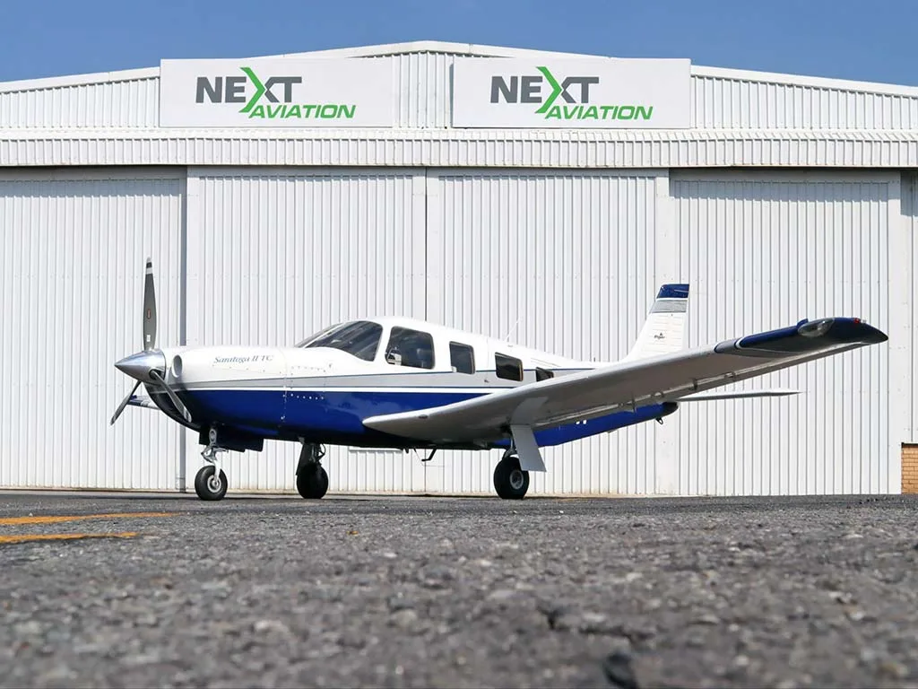 A white and blue Piper Saratoga II TC aircraft parked on an asphalt ramp in front of a hangar.