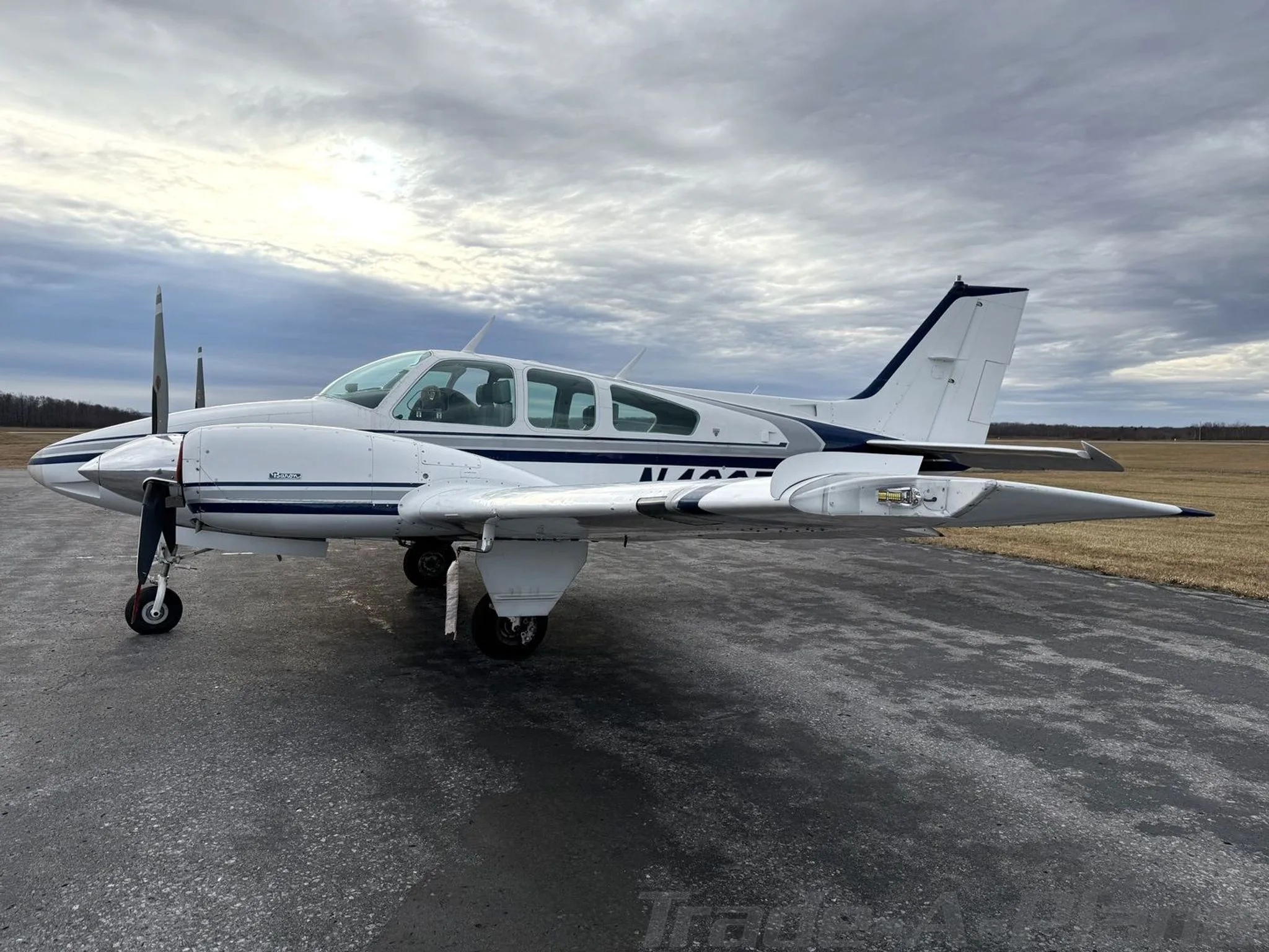 Side view of a white twin-engine aircraft with blue and grey stripes parked on an asphalt ramp under a cloudy sky.