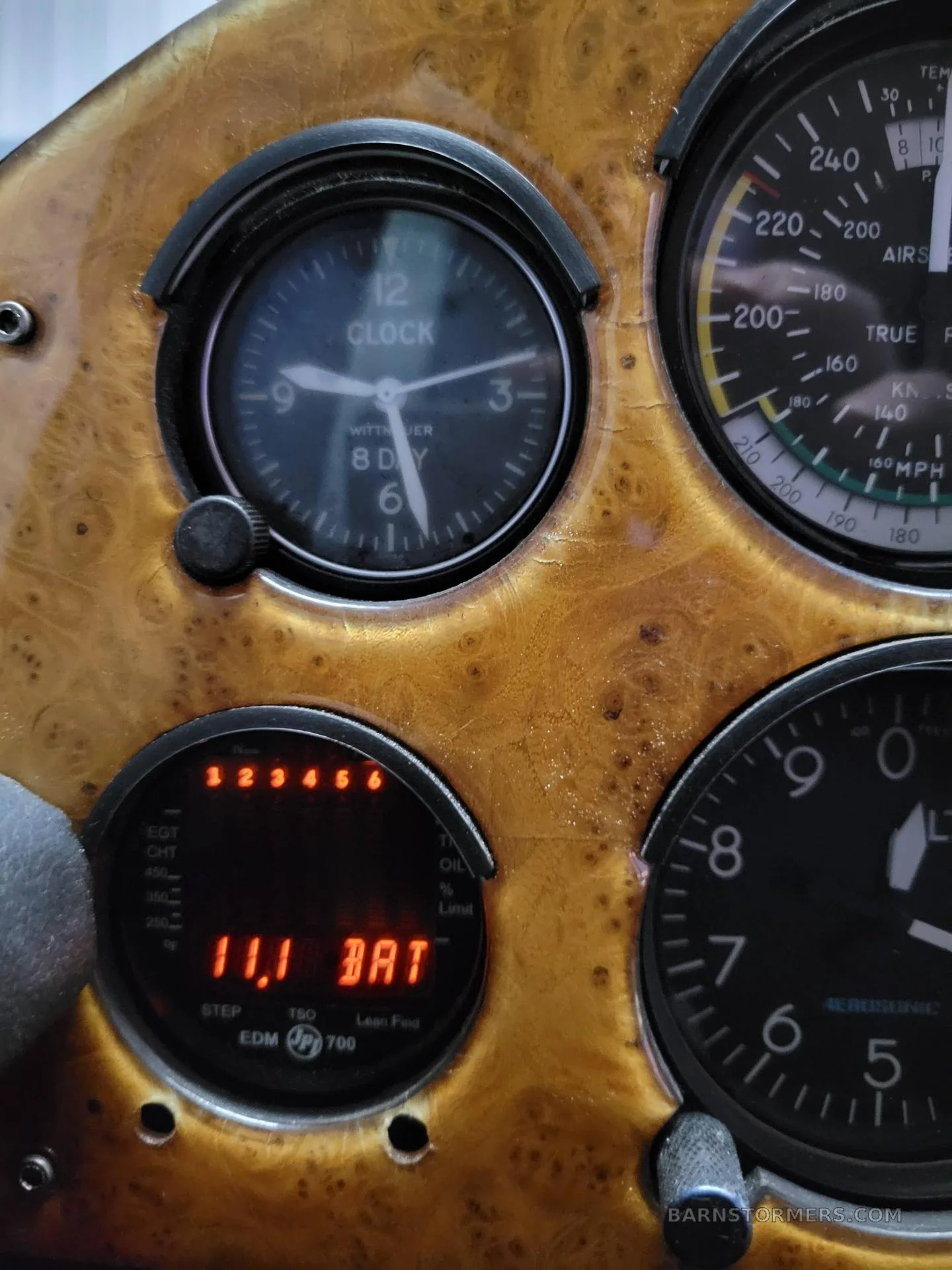 A close-up of an aircraft's wood-grain instrument panel showing a clock, airspeed indicator, and engine monitor.