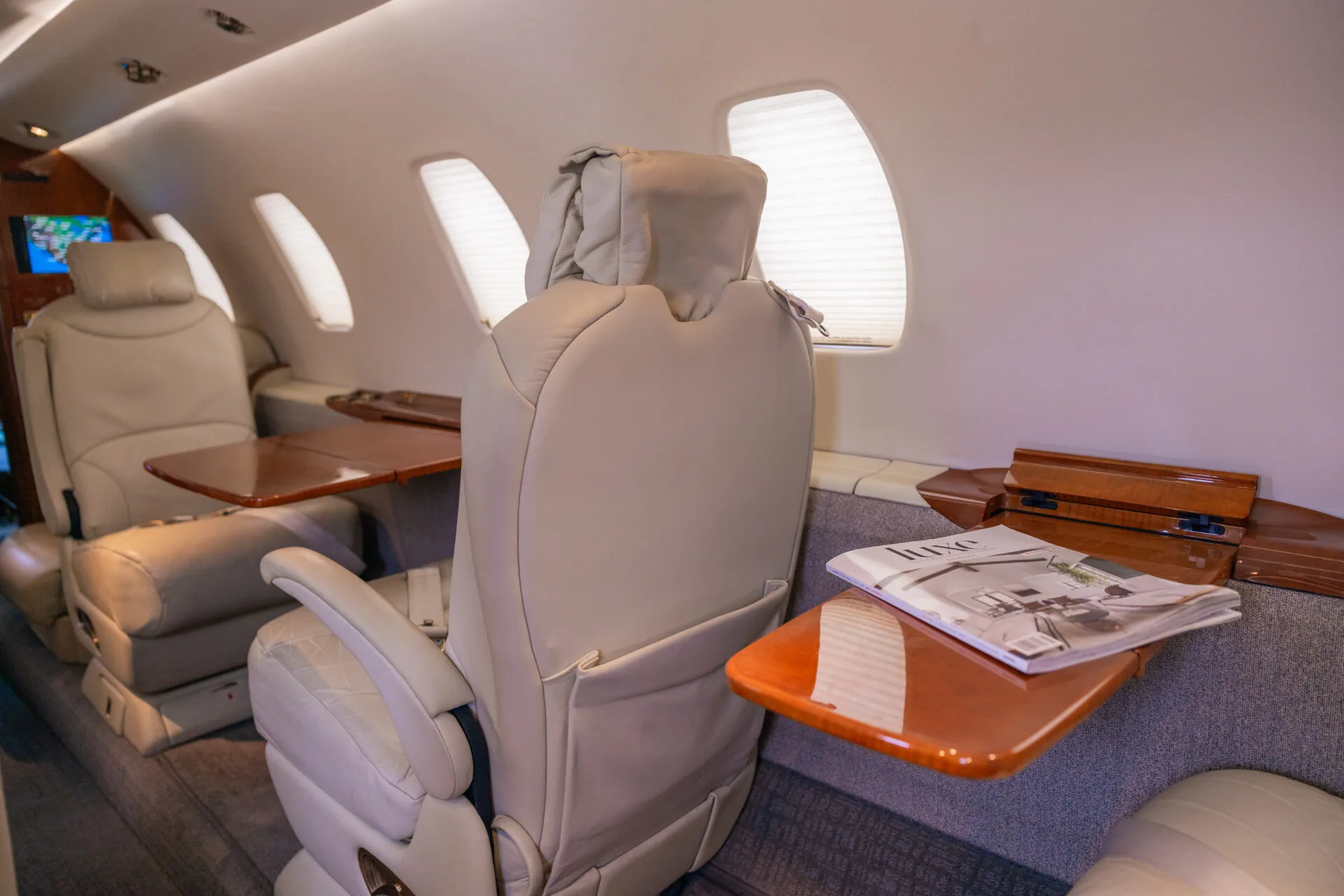 A view inside an aircraft cabin showing beige leather seats, wooden tables, and windows with shades.