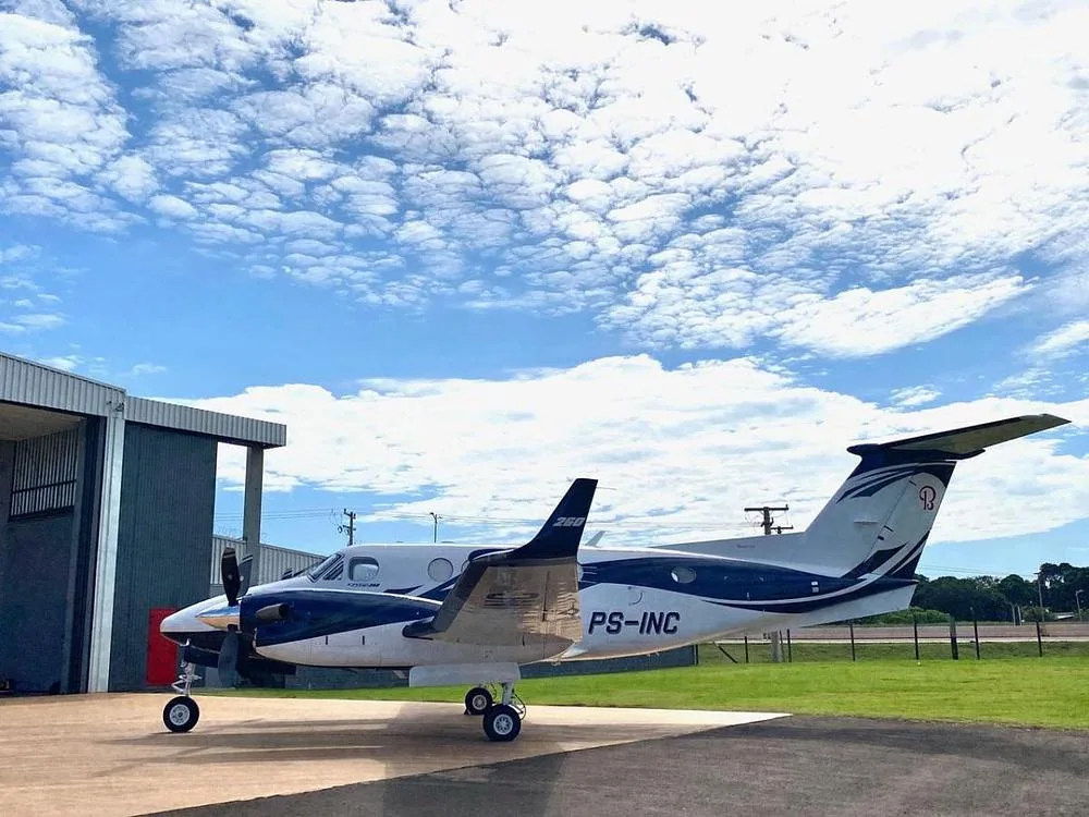 A white and blue Beechcraft King Air twin-turboprop aircraft with registration PS-INC parked on a tarmac next to a hangar.