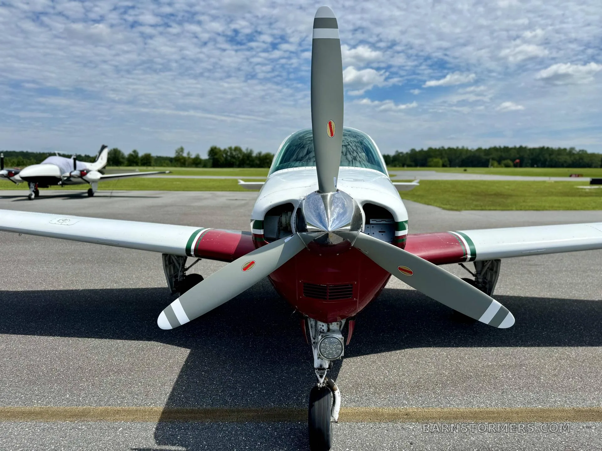 A close-up front view of the propeller and nose of a white and maroon single-engine aircraft on an airfield.