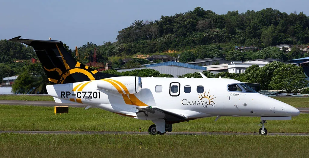 A white Embraer Phenom 300E with yellow and black Camaya Air livery on a runway with trees in the background.