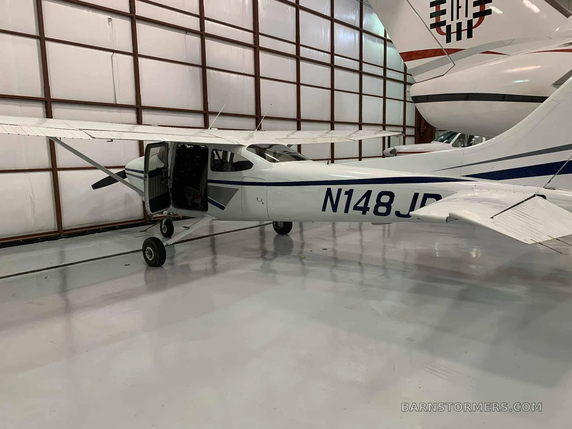 A white single-engine aircraft with blue and grey stripes, registration N148JB, parked in a hangar with its door open.