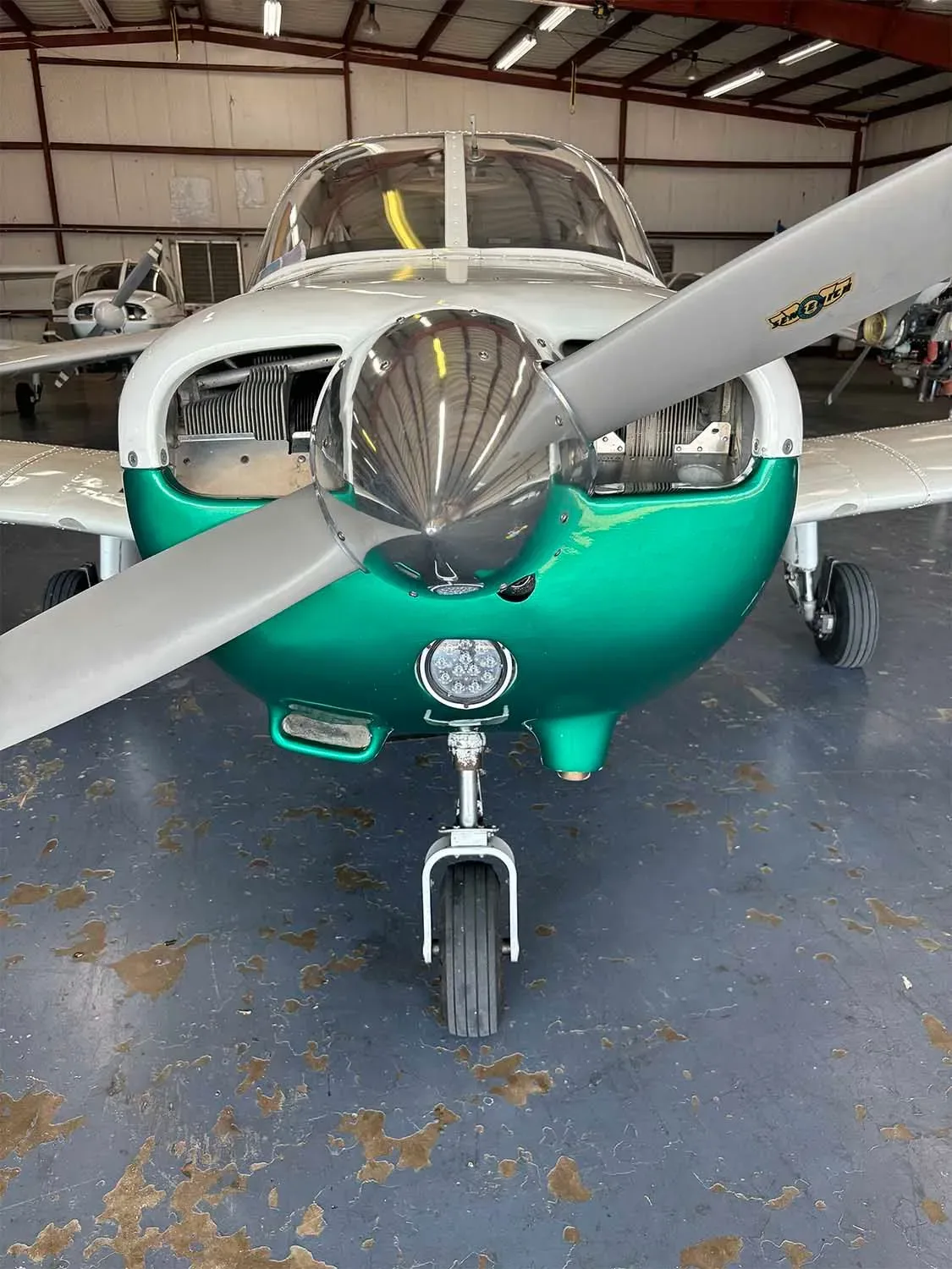 A close-up front view of a white and green single-engine aircraft with its engine cowling open in a hangar.