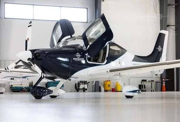 A dark blue and white single-engine aircraft with its gull-wing doors open, parked in a hangar.