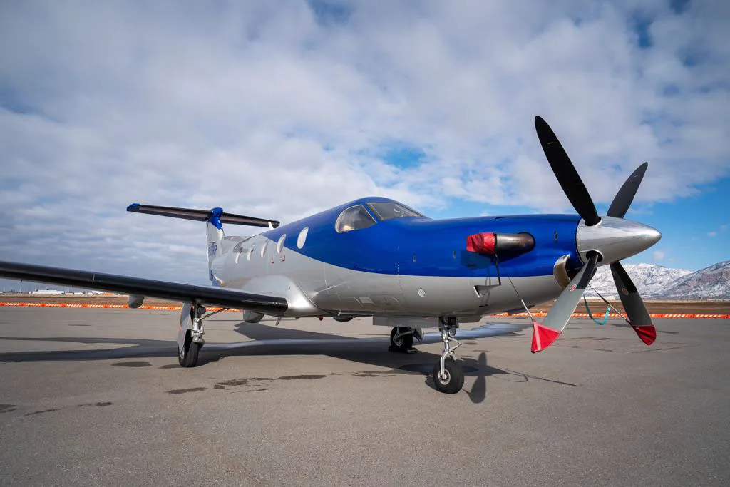 A front-side view of a blue and silver Pilatus PC-12 aircraft with its propeller blades visible, parked on an airfield under a partly cloudy sky.