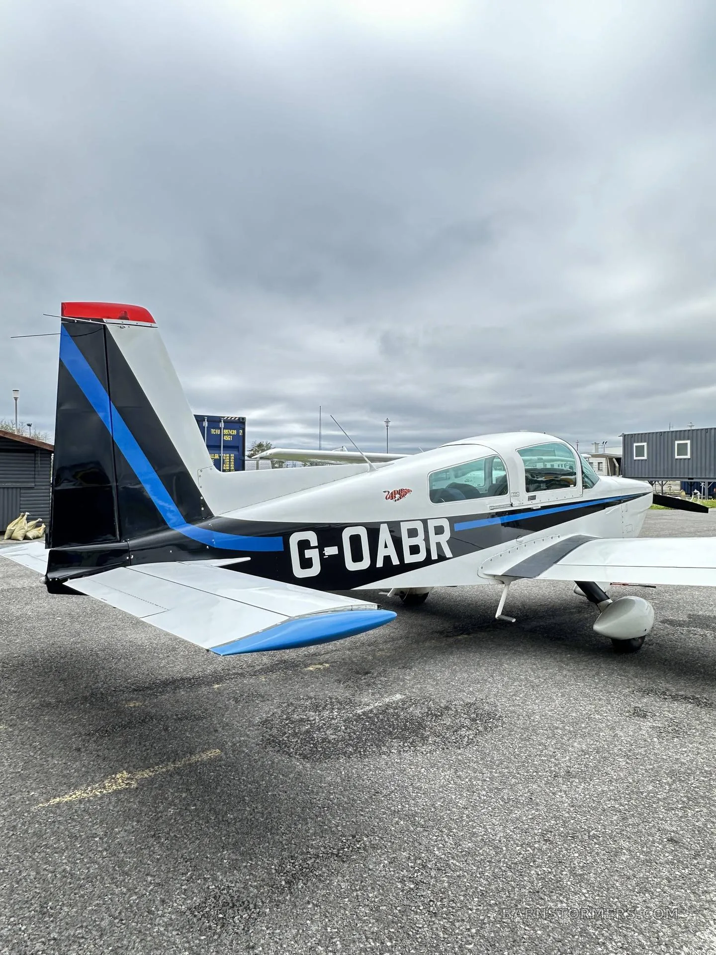 A white and blue light aircraft, registration G-OABR, parked on an asphalt apron under an overcast sky.
